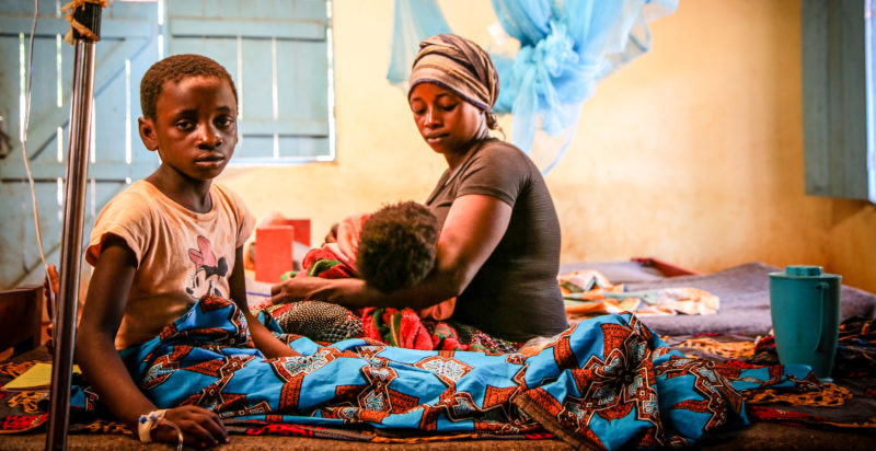 Kentia sitting upright hours after receiving an IV malaria treatment, and her mother sitting on the bed with her and her brother