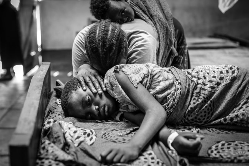 Kentia, a 9-year-old refugee in Tanzania, laying in the hospital bed as her mother mourns over her severe case of malaria 